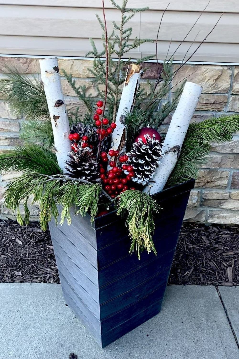 Black square wooden Christmas planter filled with white birch logs, evergreen branches, red berries and snowy pinecones