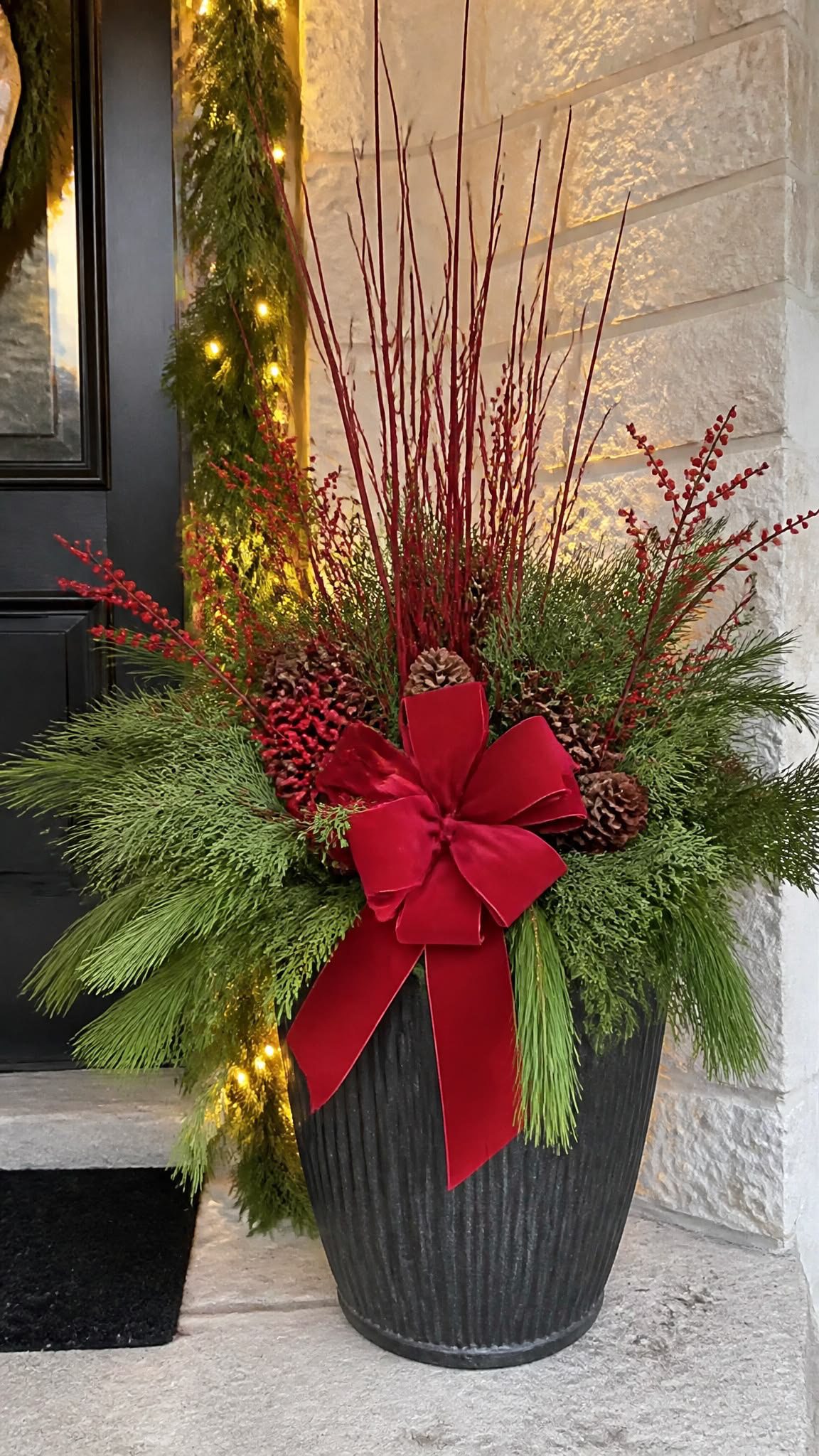 Elegant black Christmas planter with evergreen branches, tall red twigs, pinecones and large red velvet bow