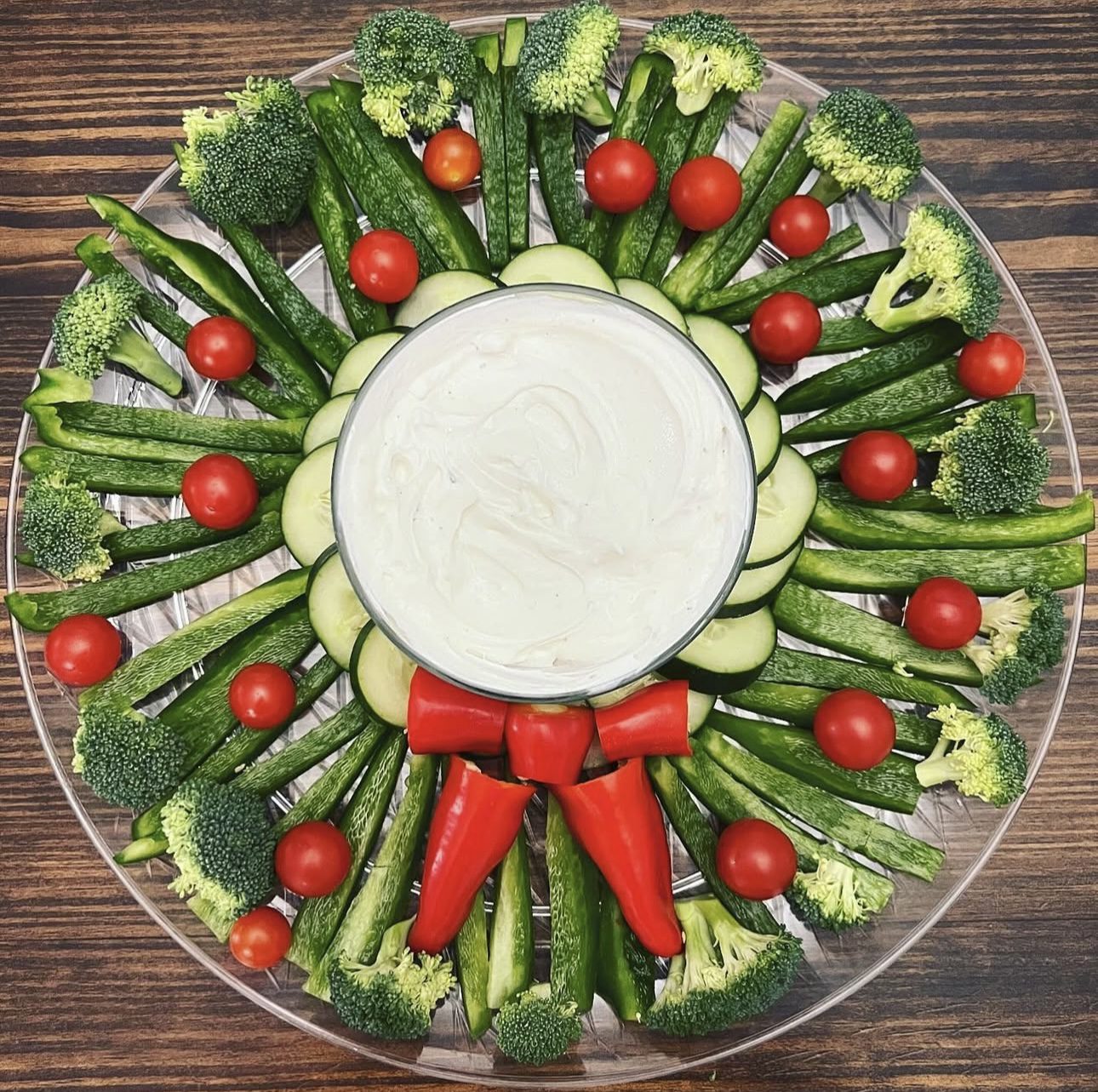 Round Christmas vegetable wreath with cucumber spears, broccoli florets, cherry tomatoes, red pepper bow and central bowl of creamy dip