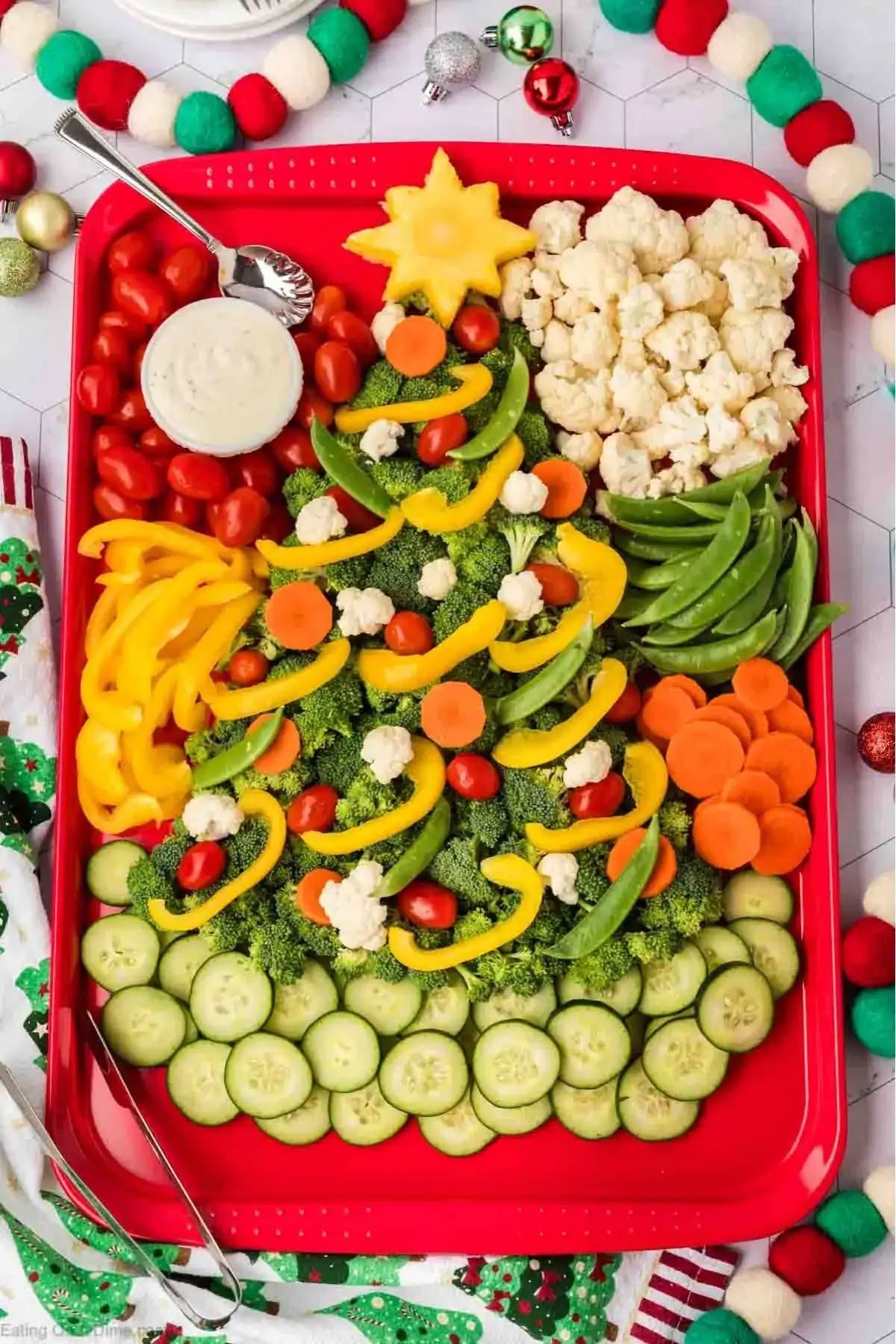Broccoli Christmas tree vegetable platter on a red tray decorated with yellow pepper garlands, carrots, tomatoes, snap peas and cauliflower