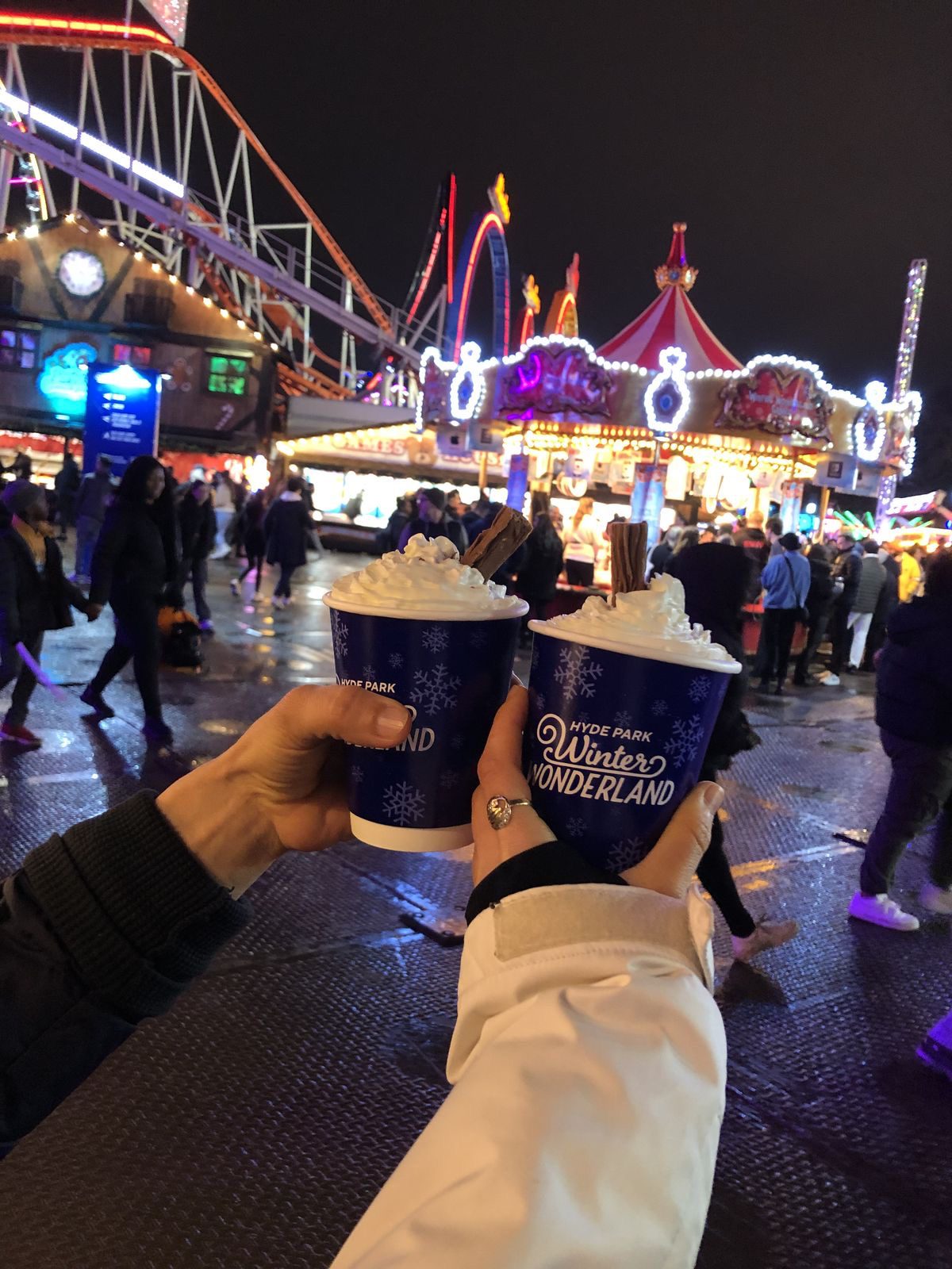 Two hot chocolates held in front of illuminated rides at a winter fair