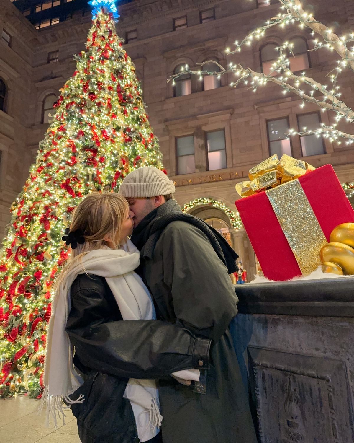 Couple sharing a kiss beside a large illuminated Christmas tree and festive decorations
