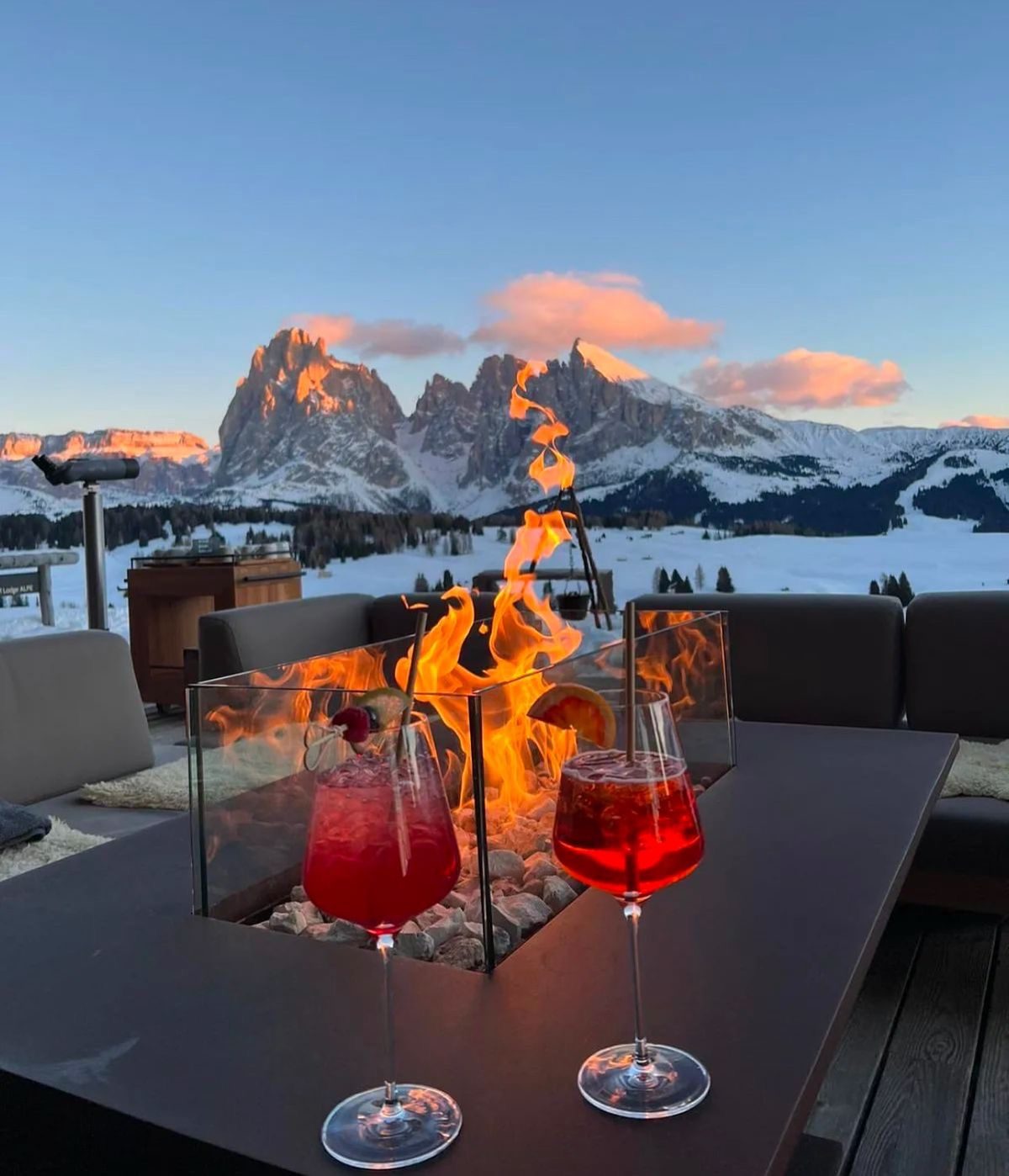 Two red drinks beside a terrace fire with snowy mountains glowing at sunset